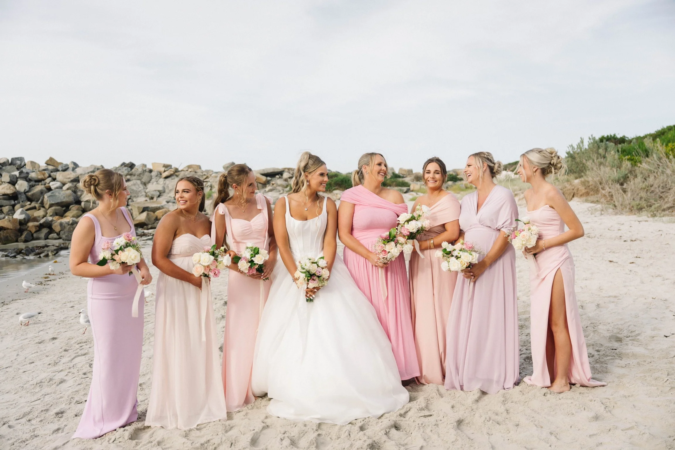A group of women for a wedding in pastel-colored dresses standing on a beach, smiling and holding bouquets of flowers, with rocks and greenery in the background.