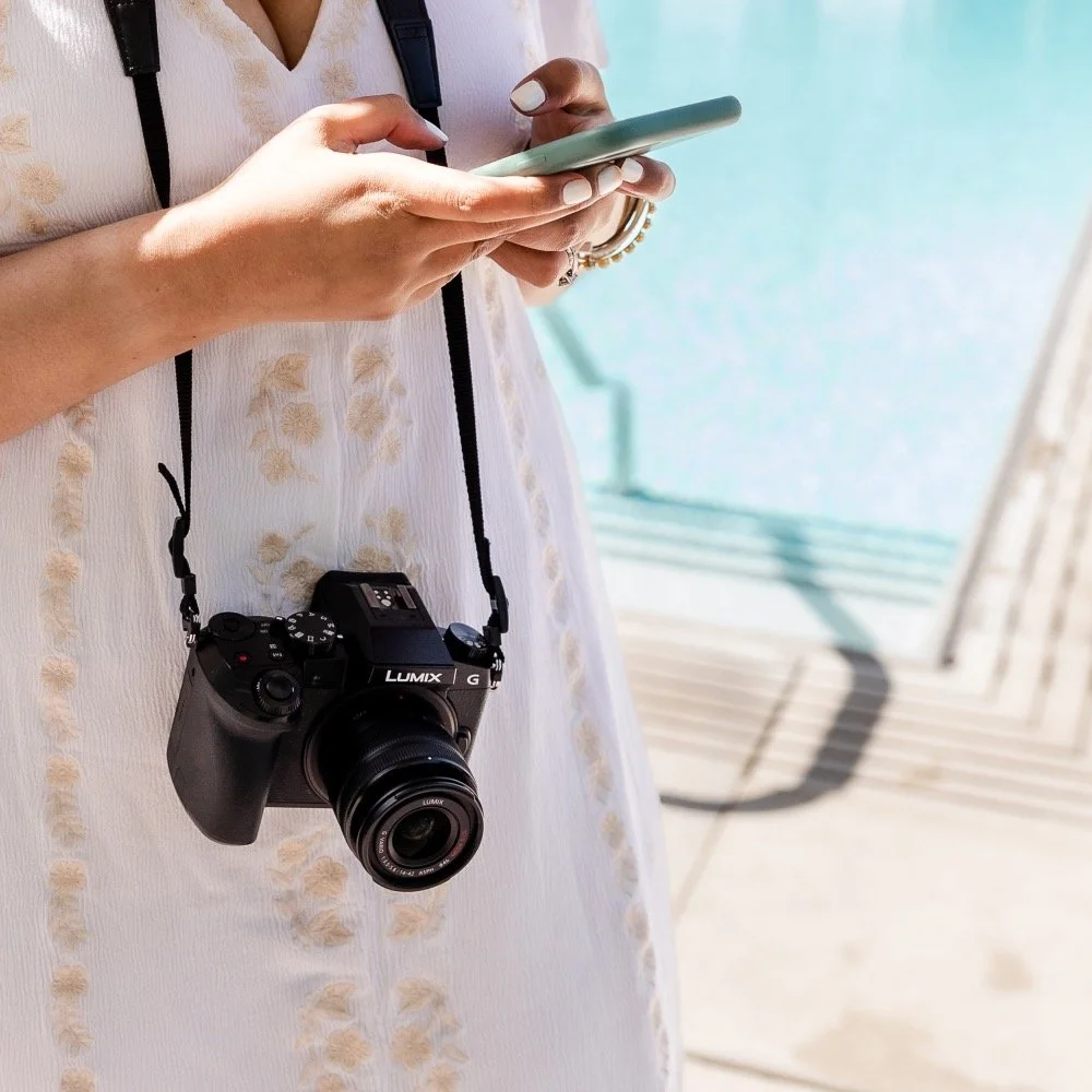 A person wearing a white, embroidered dress with a black camera hanging around their neck, holding a smartphone.