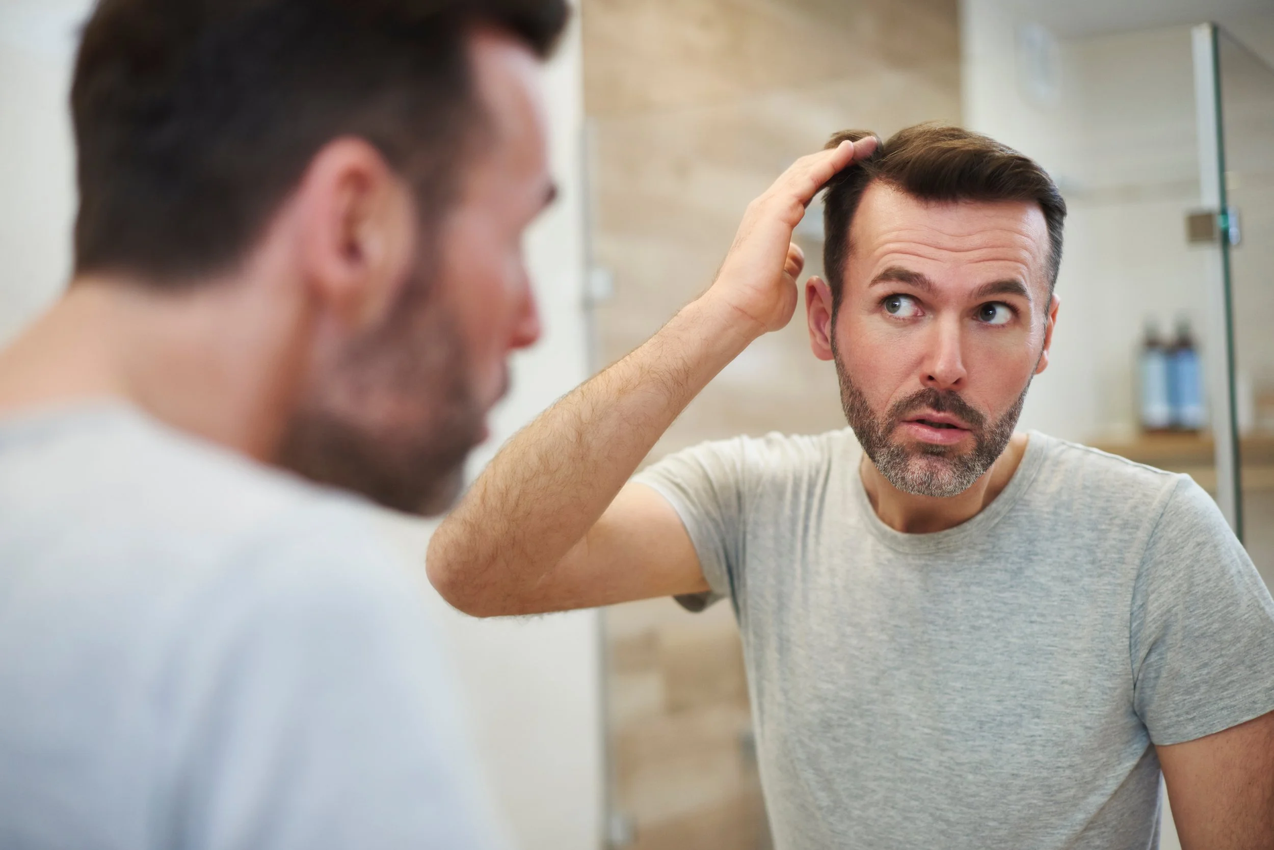A man looking in a mirror, examining his hair, with another man appearing in the foreground, slightly out of focus.
