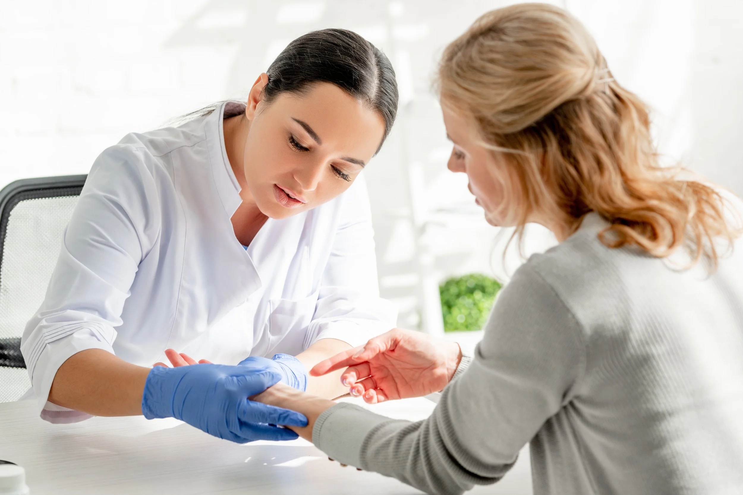 A nurse in a white uniform and blue gloves is drawing blood from a woman with red hair. The woman is sitting at a white table in a bright room, looking down at her arm.