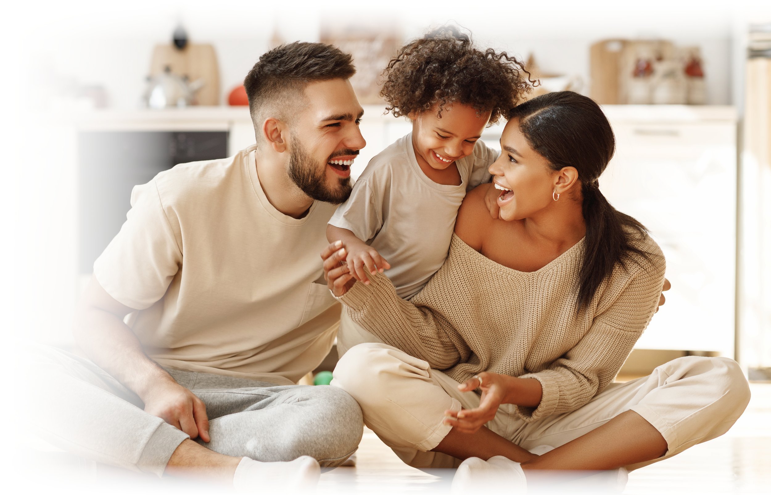 A happy family of three, parents and their young daughter, sitting on the floor in a bright kitchen, smiling and enjoying a joyful moment together.