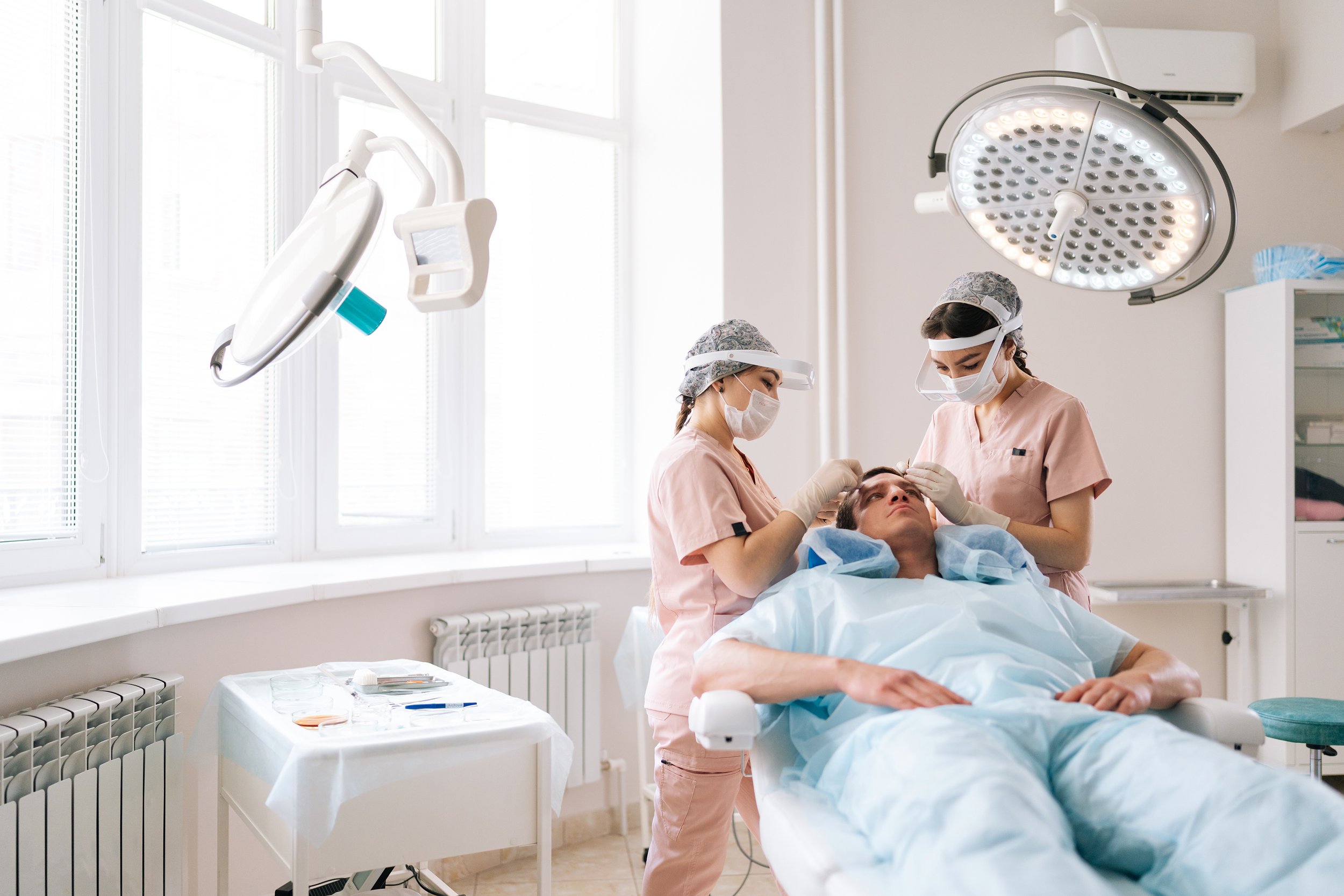 Two healthcare professionals, wearing masks and pink scrubs, are attending to a patient lying on a hospital bed. The patient appears to be receiving a procedure in a well-lit medical room with large windows and surgical lights overhead.