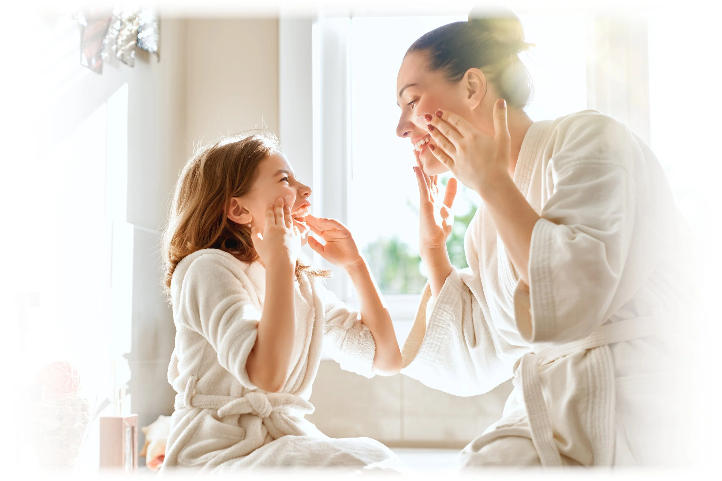 A mother and daughter laughing and playing together wearing bathrobes near a window in a bright room.