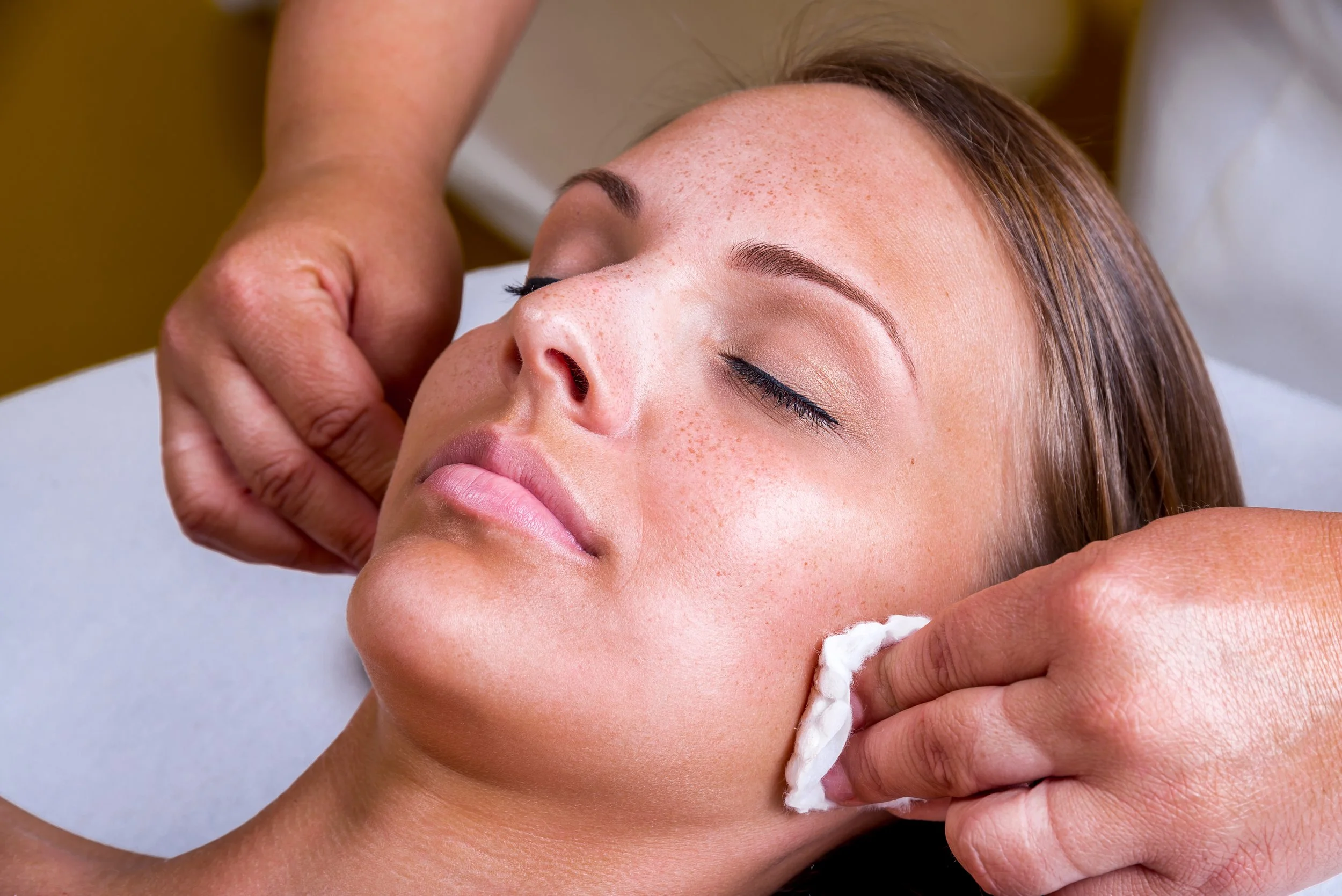 A woman receiving a facial massage or treatment, with her eyes closed and a cotton pad on her cheek.