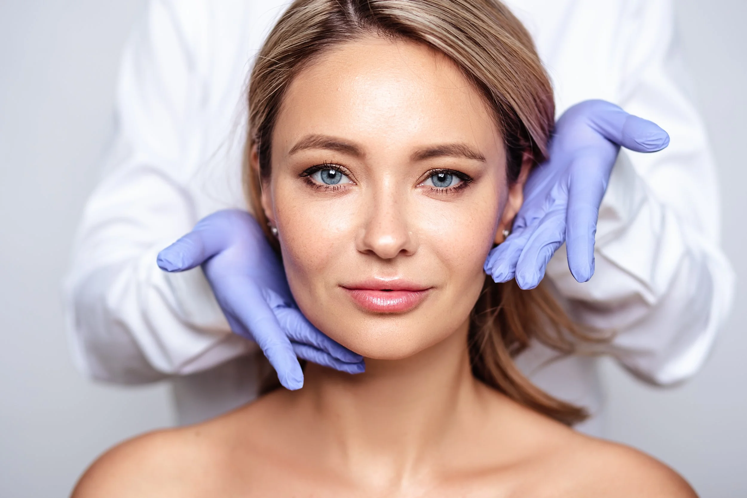 Close-up of a woman with clear skin, blue eyes, and light brown hair receiving a facial examination from a medical professional wearing a white coat and purple gloves.