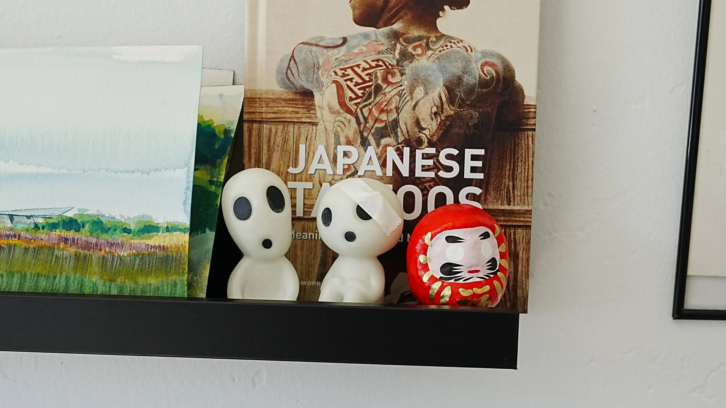 A shelf with a colorful landscape painting, two ghost-shaped figurines with surprised expressions, a Daruma doll with a cat face, and a book titled 'Japanese Tattoos' featuring a tattooed person.