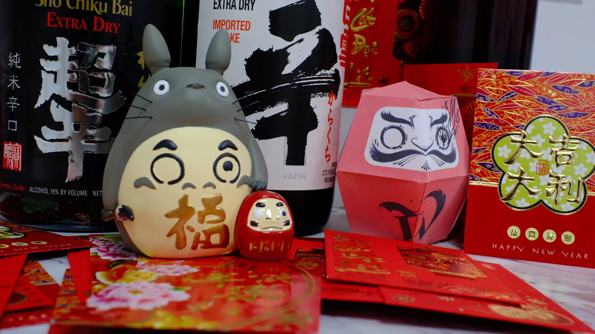 Assorted Chinese New Year decorations and figurines, including a Totoro figure, Daruma doll, and red envelopes, with traditional Chinese characters and symbols.