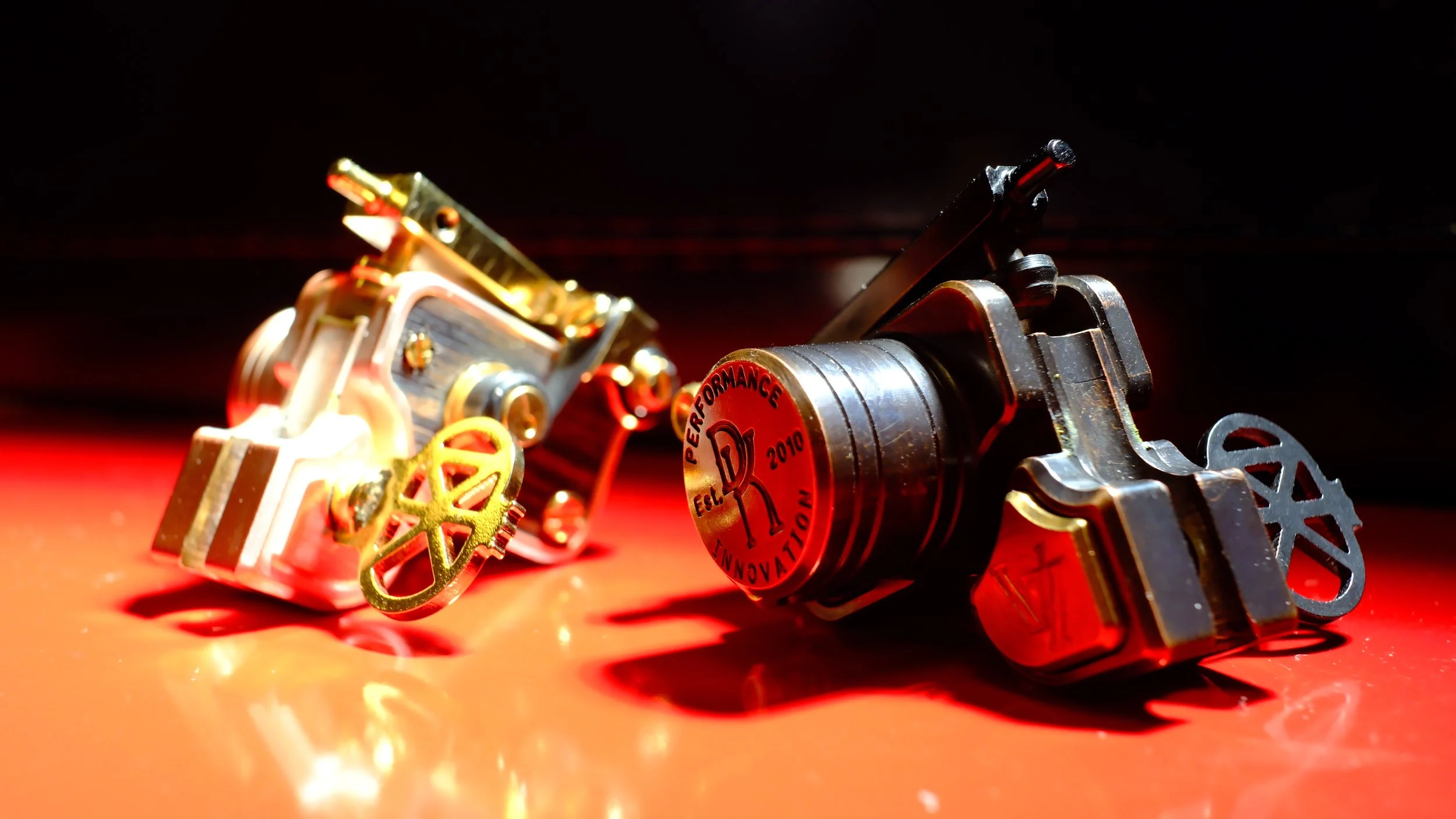 Close-up of two small mechanical parts on a red surface, with the right one featuring a round red label that says "PERFORMANCE D 2010" and a stylized "R" logo, both with gold and metal components.