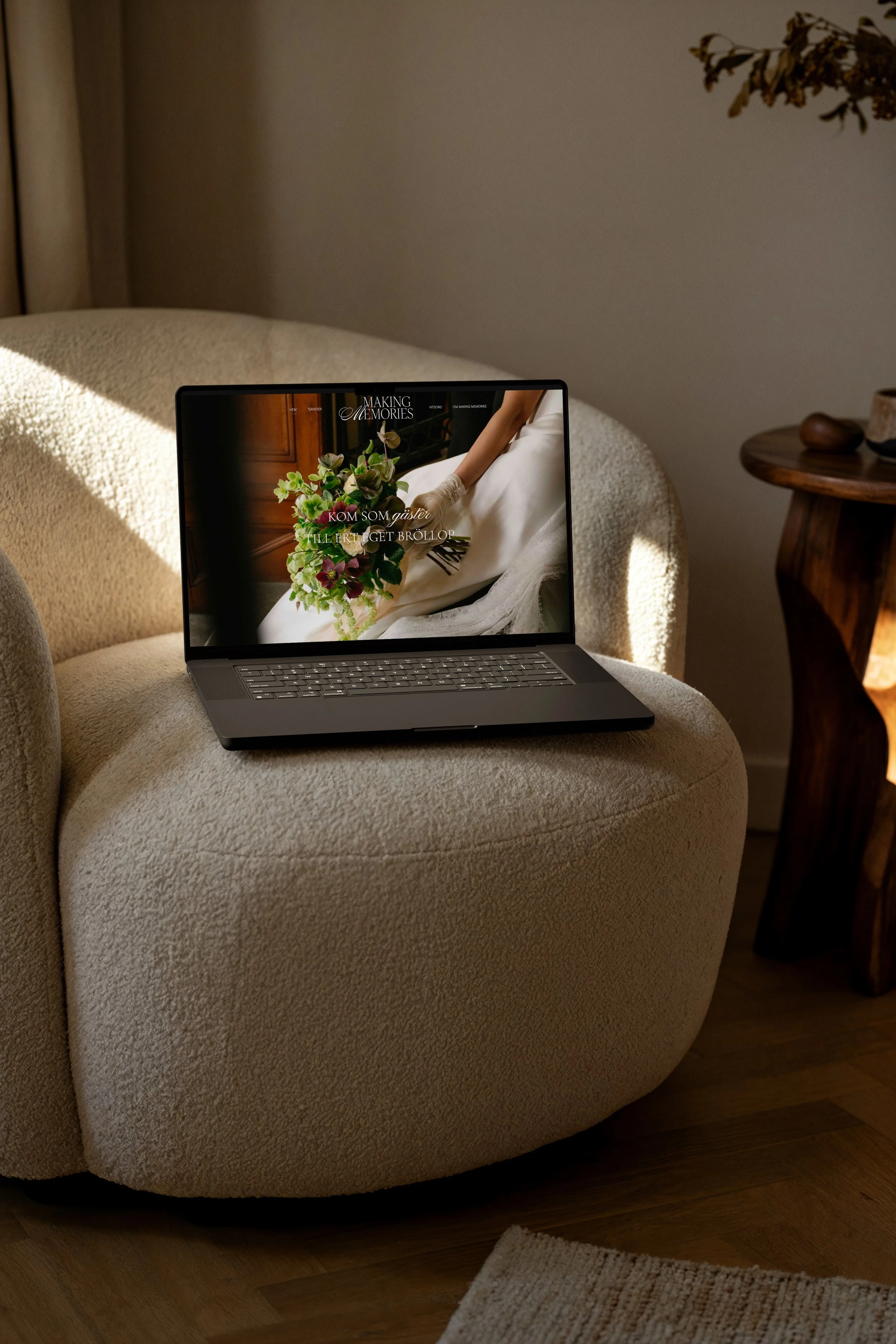 A beige sofa with a laptop on it displaying a wedding website, a wooden side table with decorative objects, and a hardwood floor with a rug.