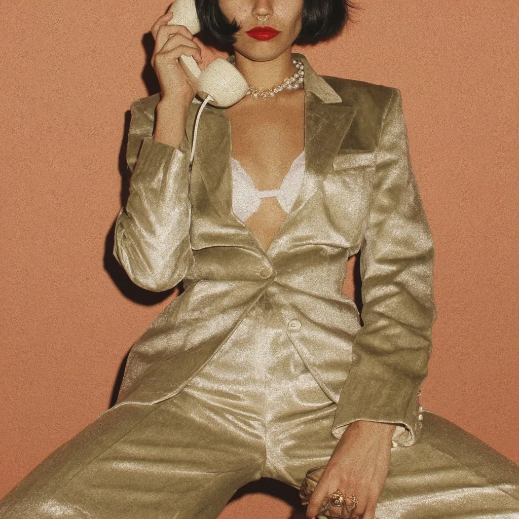 A woman in a shiny gold blazer and matching pants, holding a vintage white telephone to her ear, with a pearl necklace and bright red lipstick, sitting against a peach-colored background.