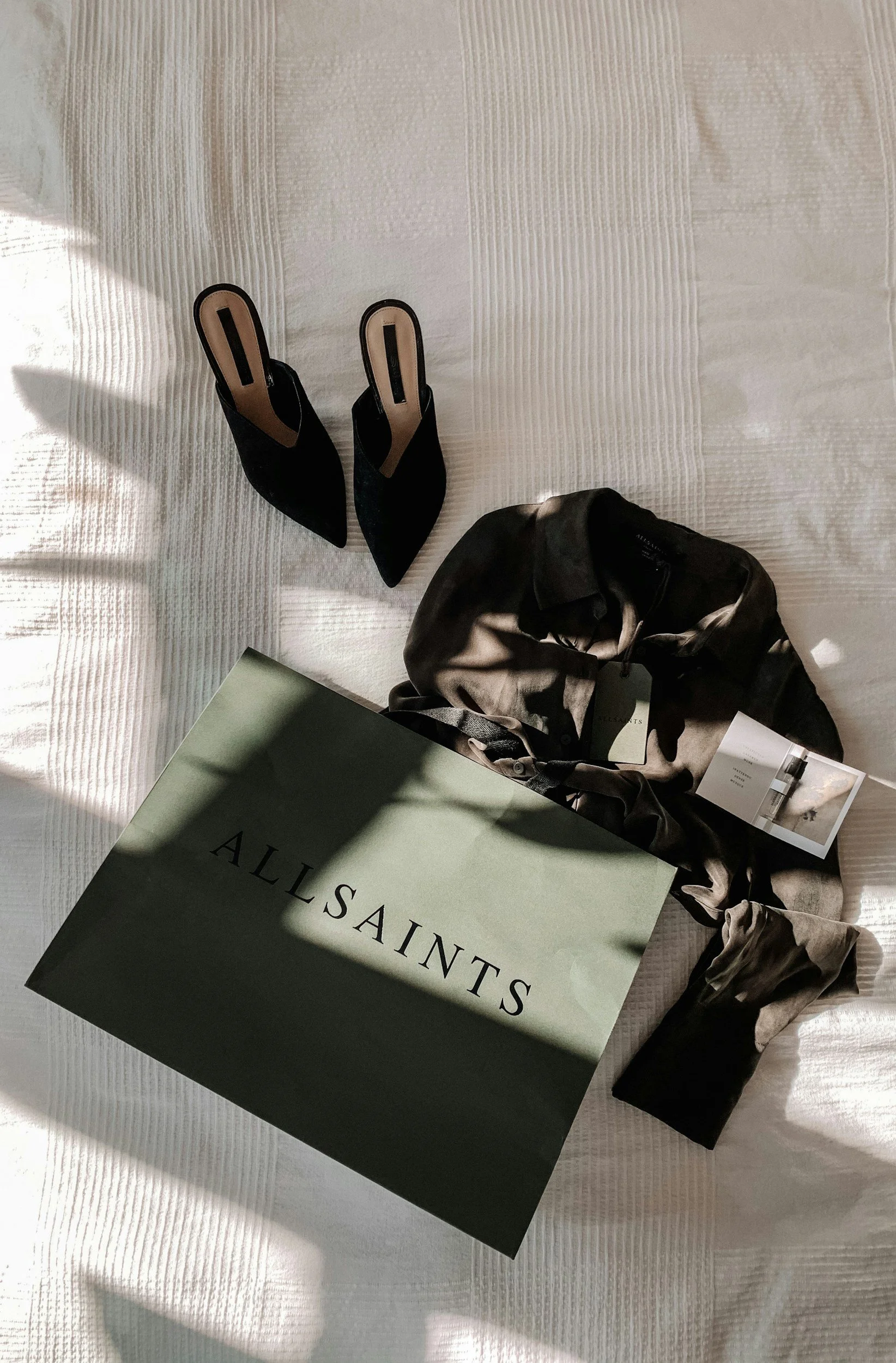 A photo of a shopping bag from AllSaints, a pair of black high-heeled shoes, and a silk shirt with tags, all placed on a white textured bedspread.