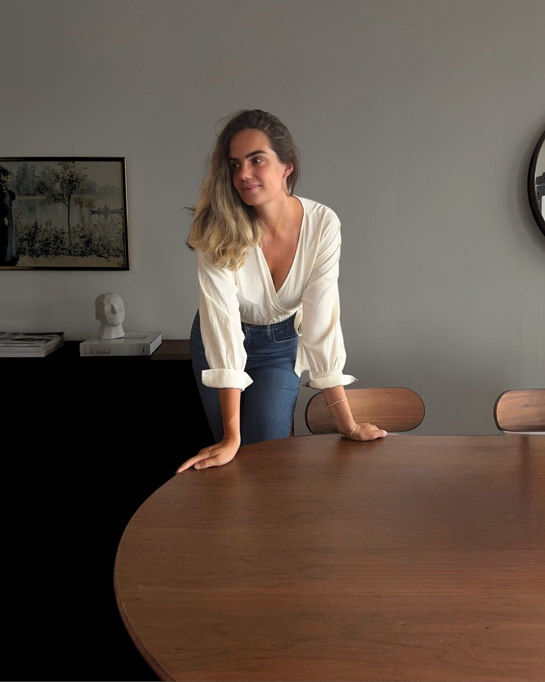 A woman with shoulder-length blonde hair leaning on a brown wooden table in a modern room, wearing a white blouse with rolled-up sleeves and dark jeans, looking to her left.