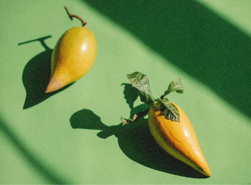 A yellow pear-shaped fruit and a yellow bell-shaped vegetable with green leaves on a green surface, casting shadows.