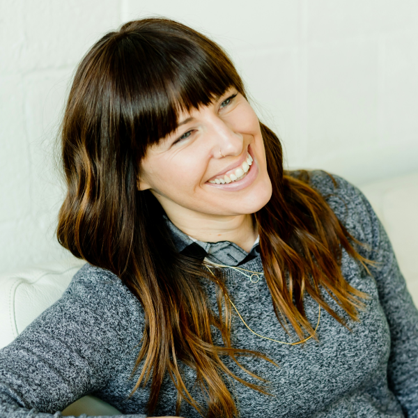 A woman with long brown hair and bangs smiling and sitting on a white couch in front of a white wall.