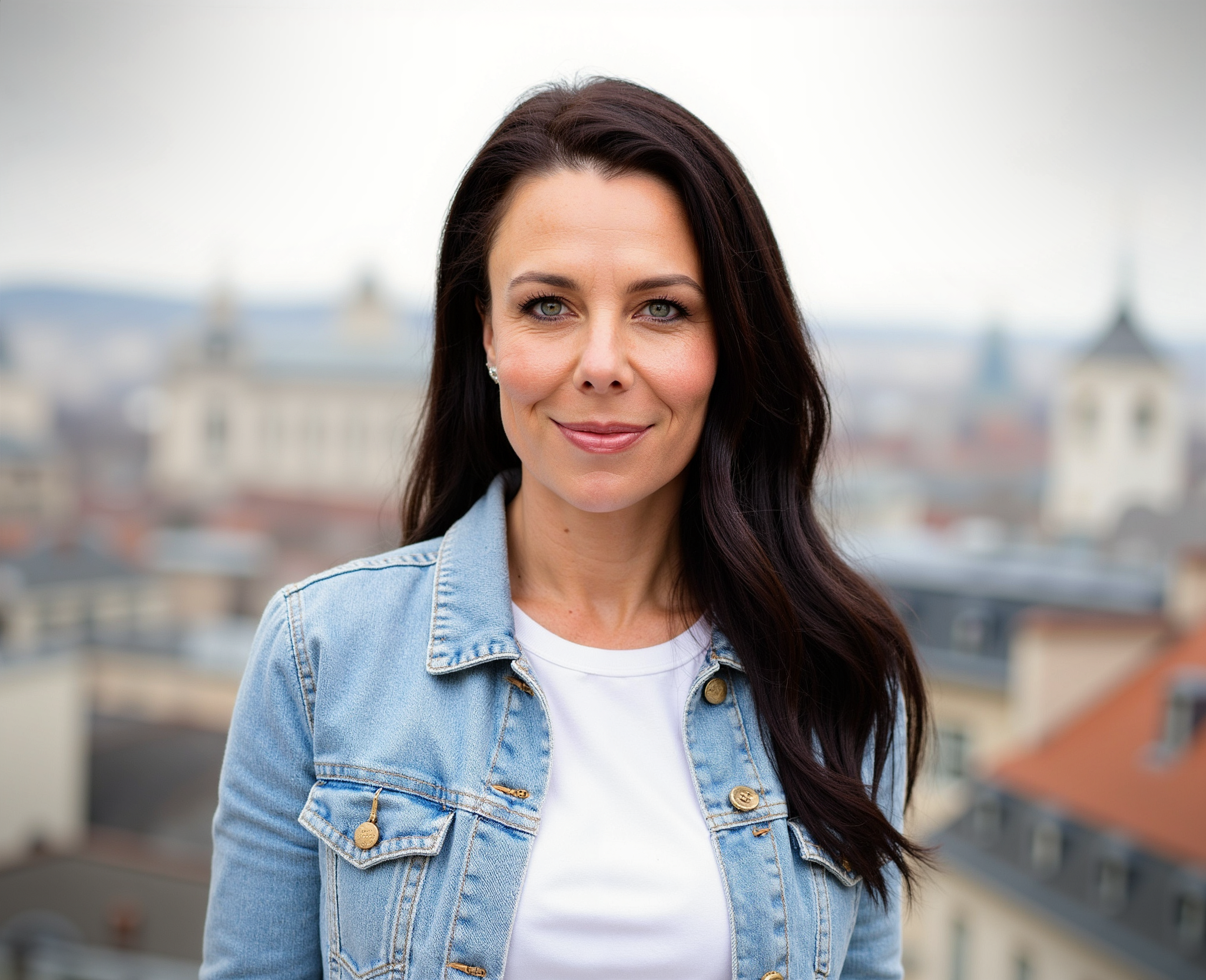 A woman with dark brown hair and blue eyes wearing a light denim jacket and white shirt, standing outdoors in front of a cityscape with buildings and rooftops.