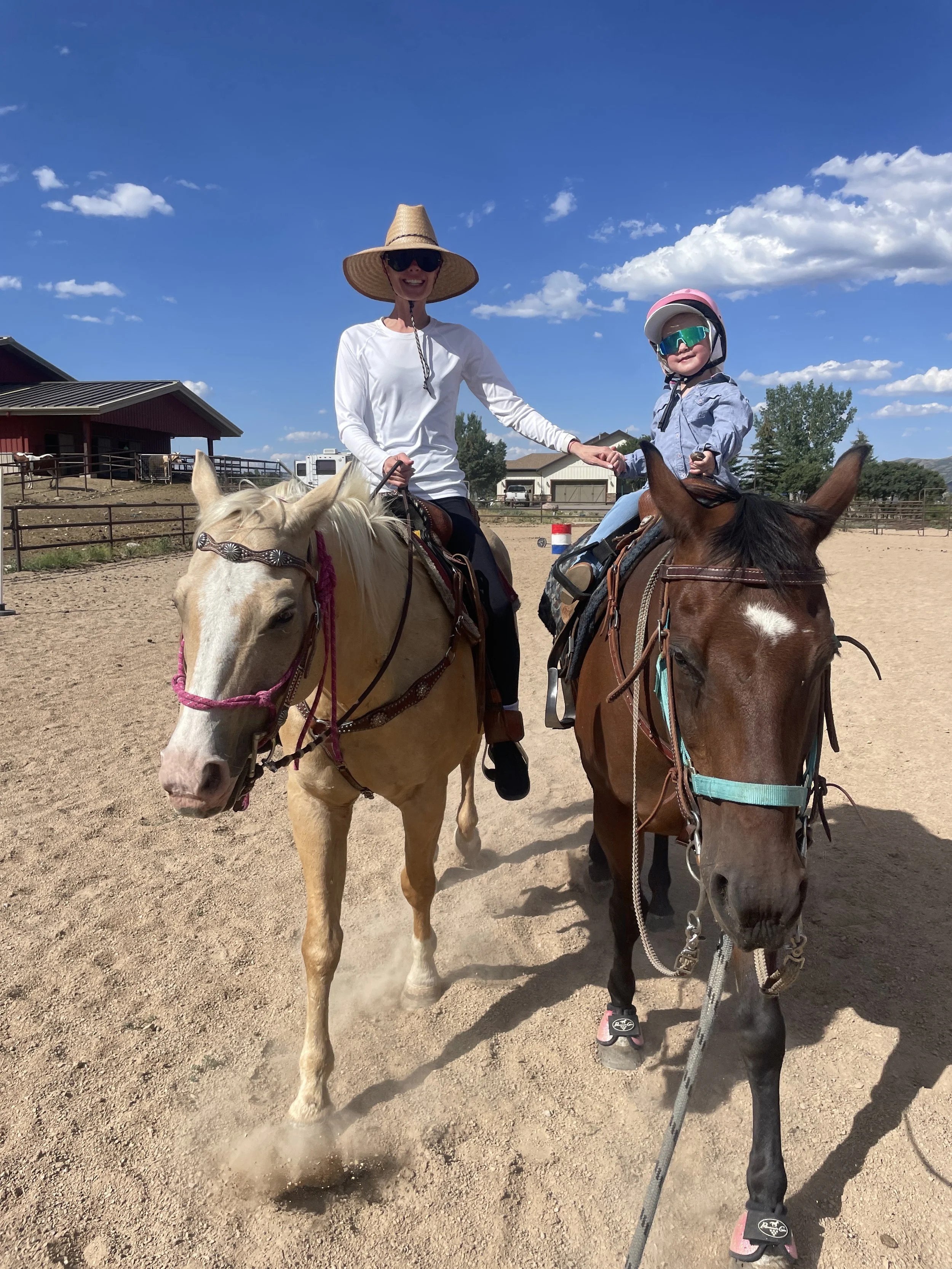 A woman and a young girl riding horses in an outdoor riding arena on a sunny day. The woman is wearing a wide-brimmed straw hat and sunglasses, and the girl is wearing a helmet and sunglasses. The arena is surrounded by rail fences and some buildings in the background.