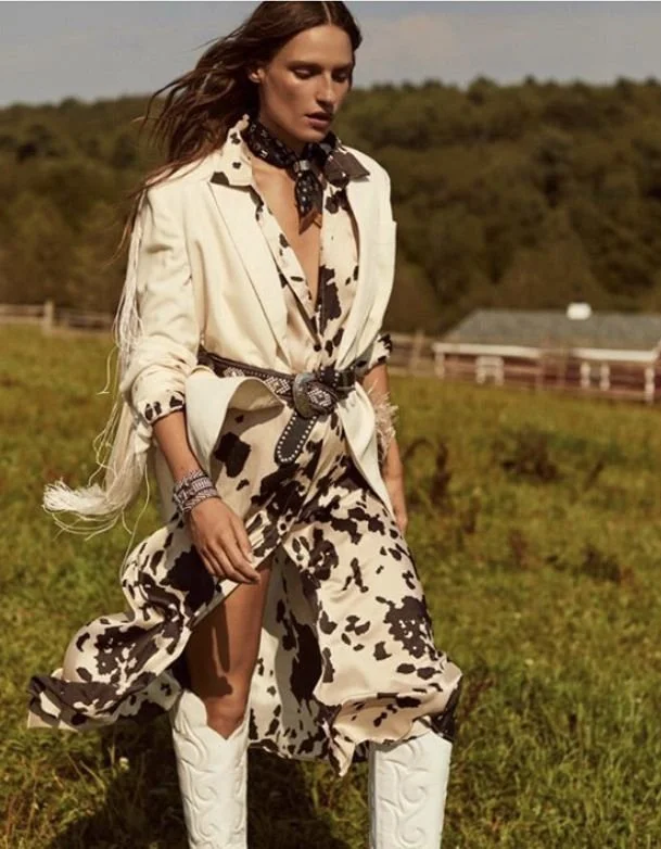 Woman in Western-style outfit with cow print patterns, cowboy boots, and a bandana, standing outdoors in a grassy field with trees and a barn in the background.