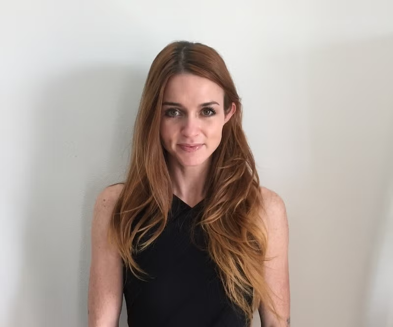 A woman with long red hair and a black sleeveless top standing against a plain white wall.