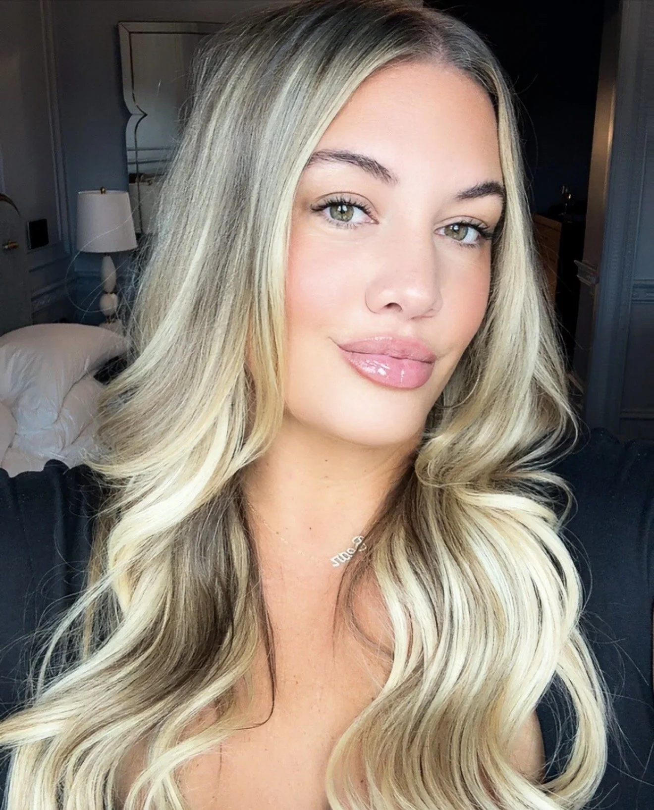 Close-up of a woman with long blonde hair, green eyes, and light makeup, wearing a black top, indoors in a well-lit room.