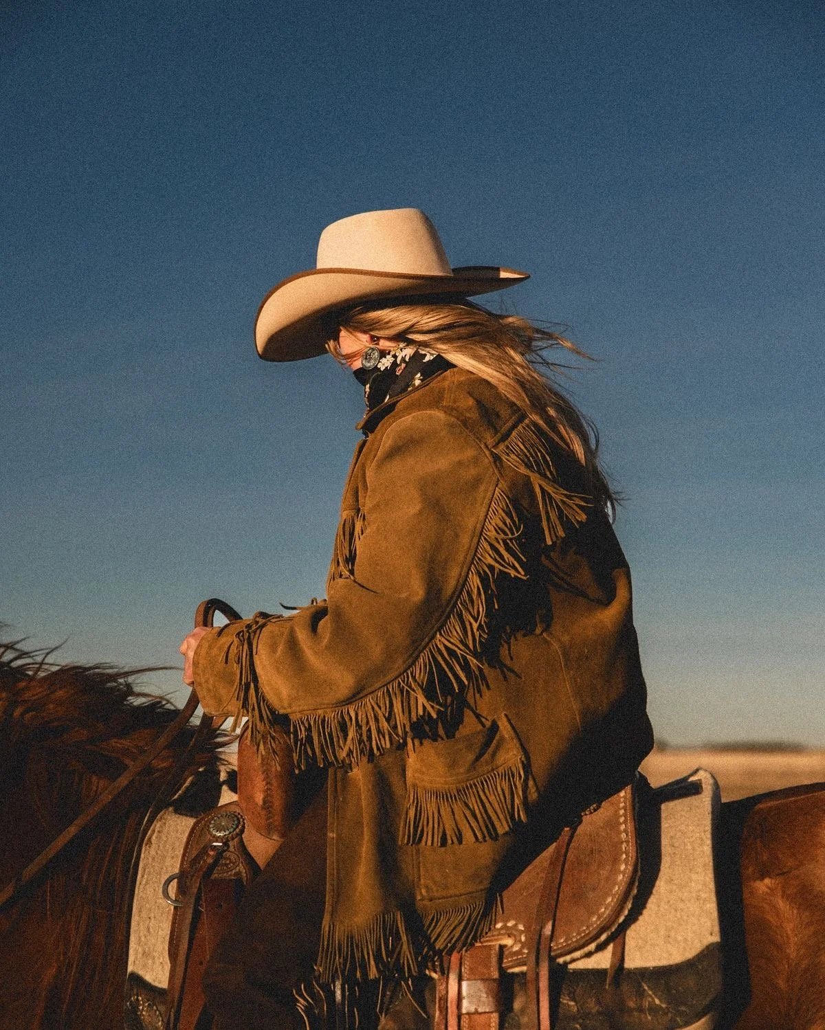 Woman wearing a cowboy hat, bandana, and fringed jacket riding a horse in a desert landscape during sunset.