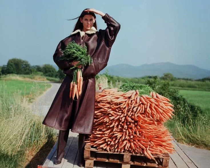 A woman standing outdoors on a farm path, holding a bunch of carrots and spinach, with a large pile of carrots on a wooden cart next to her.