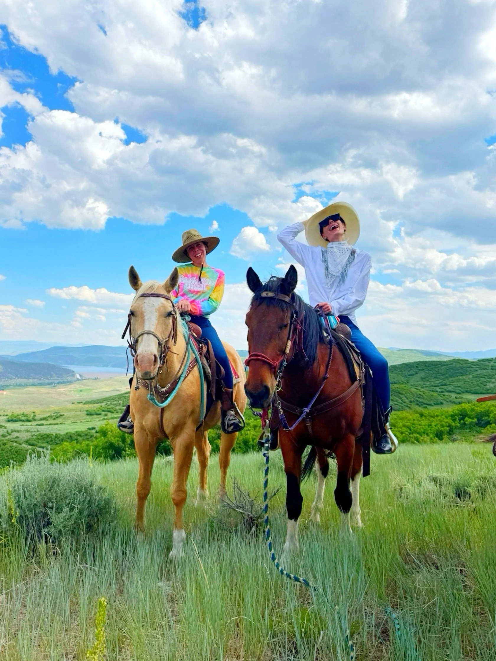 Two women riding horses in a grassy field under a partly cloudy sky, one wearing a colorful tie-dye shirt and straw hat, the other wearing a white shirt and large sun hat, both smiling and enjoying the outdoors.