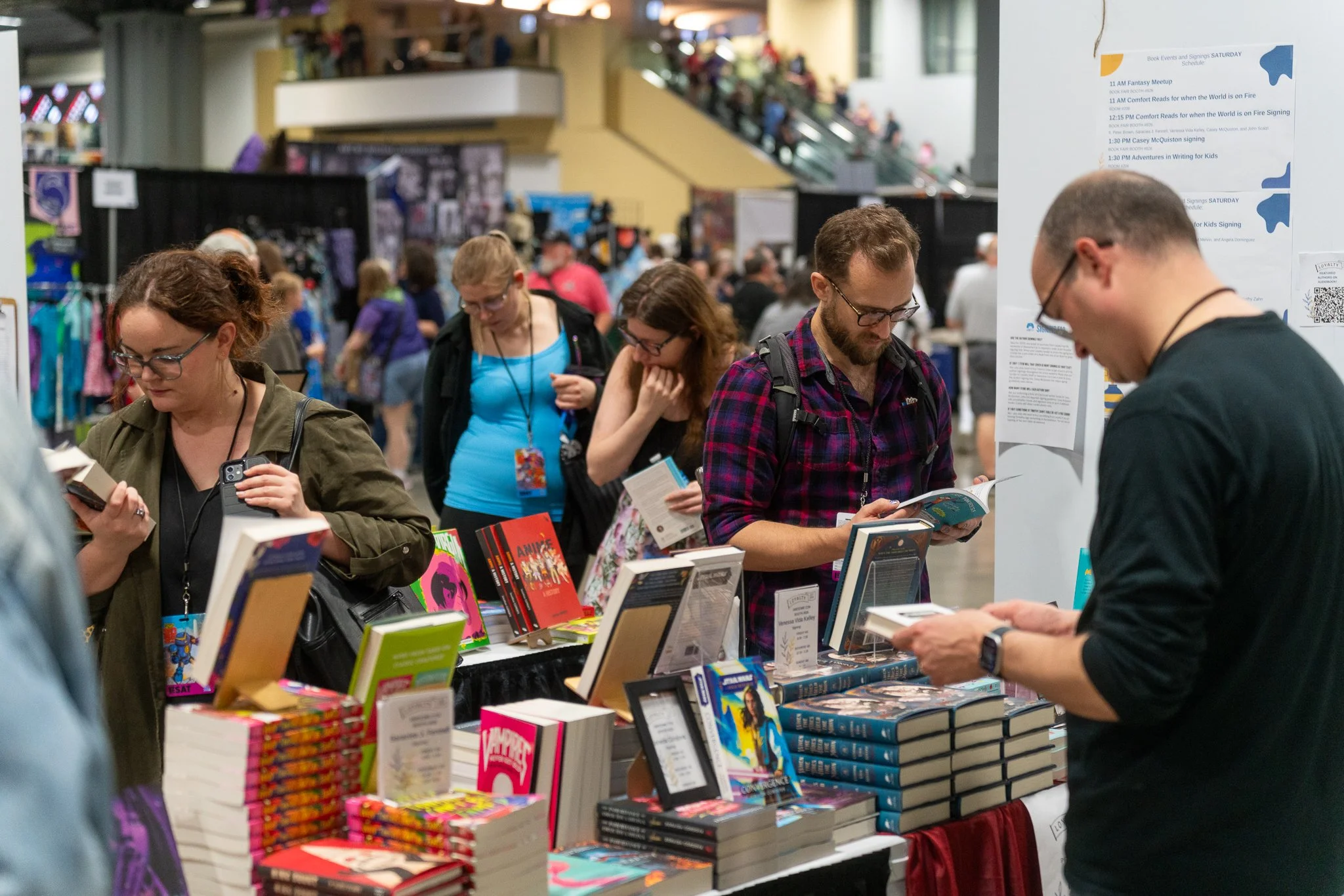 People browsing books at a convention or book fair, with tables of books and a busy background of attendees and booths.