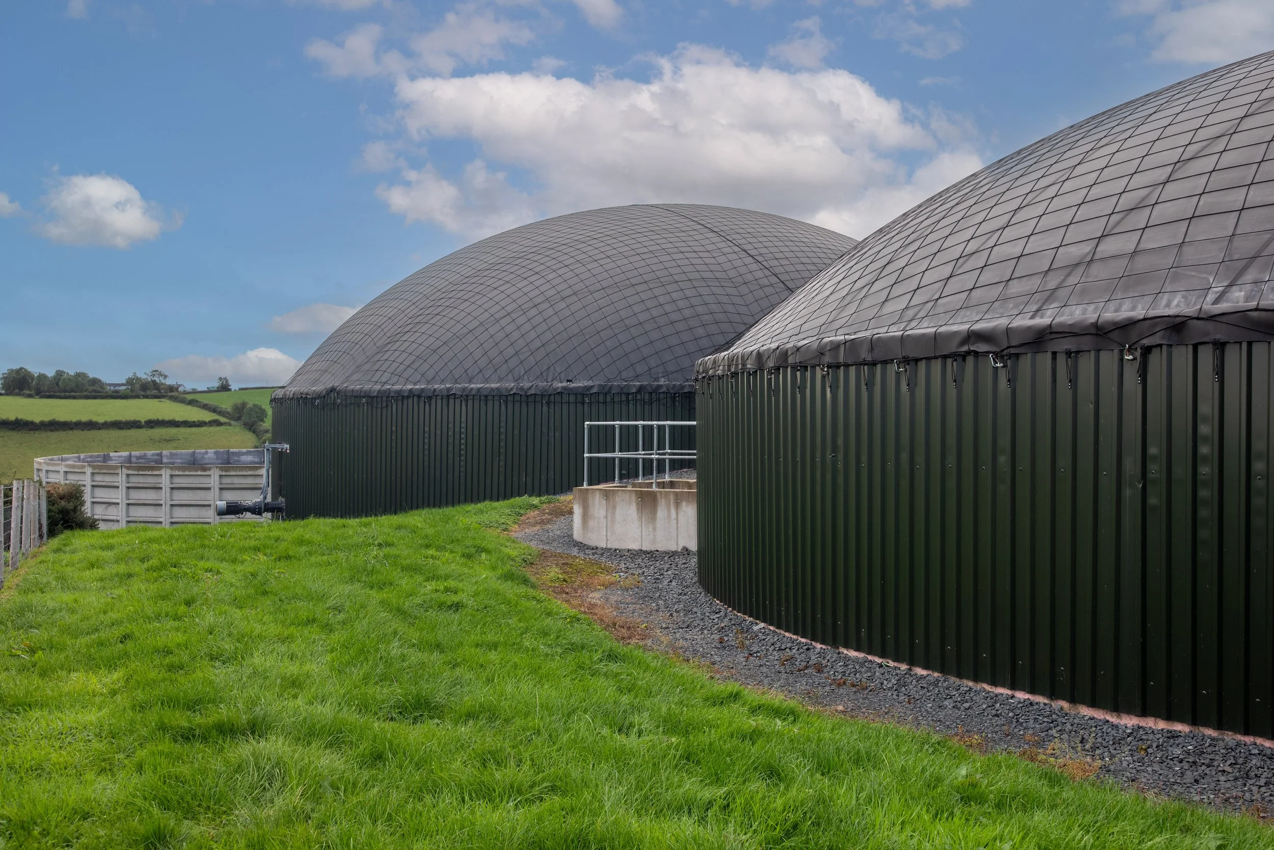 Two large, semi-circular agricultural silos with green metal exteriors and domed tops, situated on a grassy hillside under a partly cloudy sky.