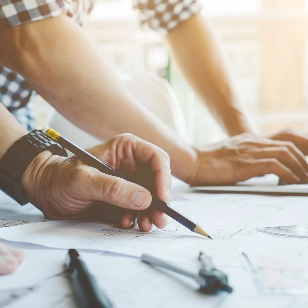Close-up of a person drawing sketches or plans on paper with a pencil, with other drawing tools on a table, in a bright environment.