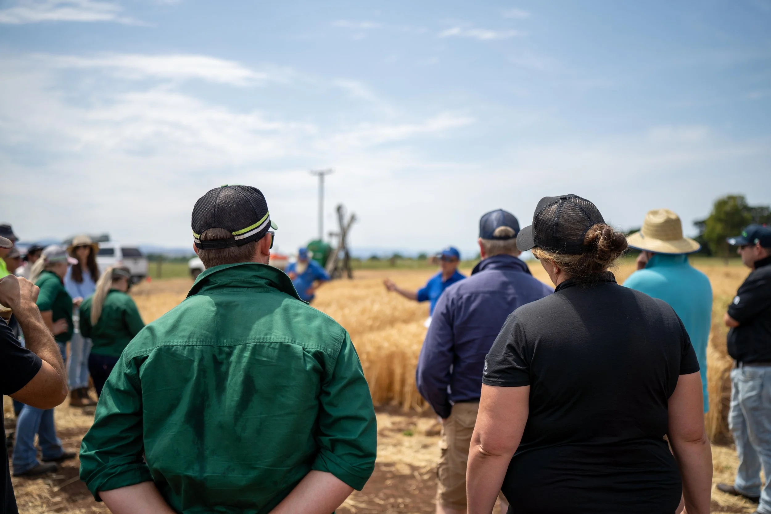 A group of people gather outdoors in a field, listening to a man in a blue shirt giving a talk or presentation amidst tall wheat or grass under a partly cloudy sky.