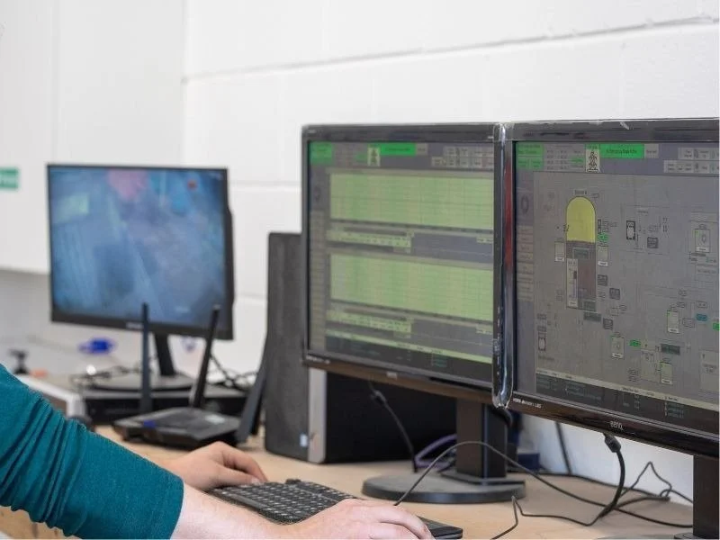 Two computer monitors displaying security footage and building layout diagrams, with a person seated at a desk operating a keyboard.