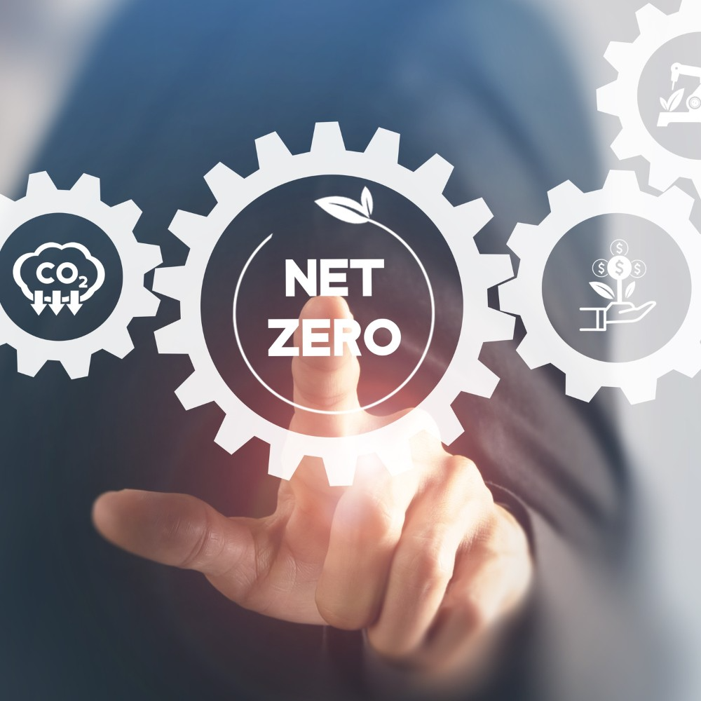A person in a suit touching a virtual interface with gears symbolizing environmental sustainability. The central gear reads 'NET ZERO' with a leaf icon, surrounded by icons representing reducing CO2 emissions and promoting green investments.