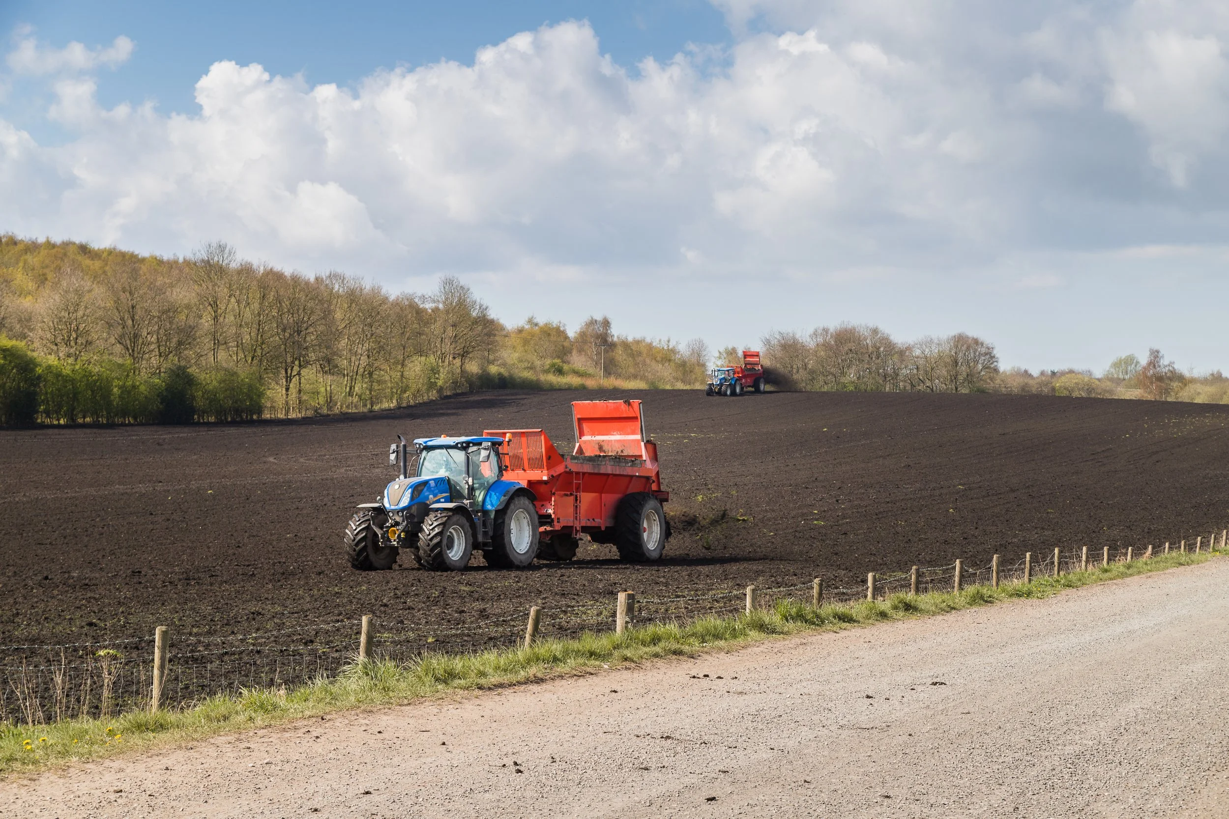Two tractors working on a dark, freshly tilled farm field with a dirt road in the foreground, a line of trees in the background, and a partly cloudy sky overhead.