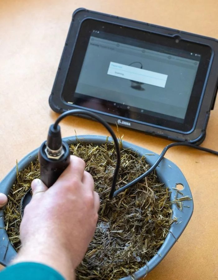 A person testing soil on a patchy, dried plant material using a handheld device connected to a tablet screen displaying a measurement interface.
