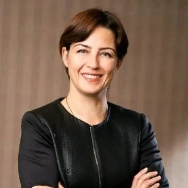 A woman with short brown hair, wearing a black jacket, smiling and crossing her arms, standing indoors against a neutral background.
