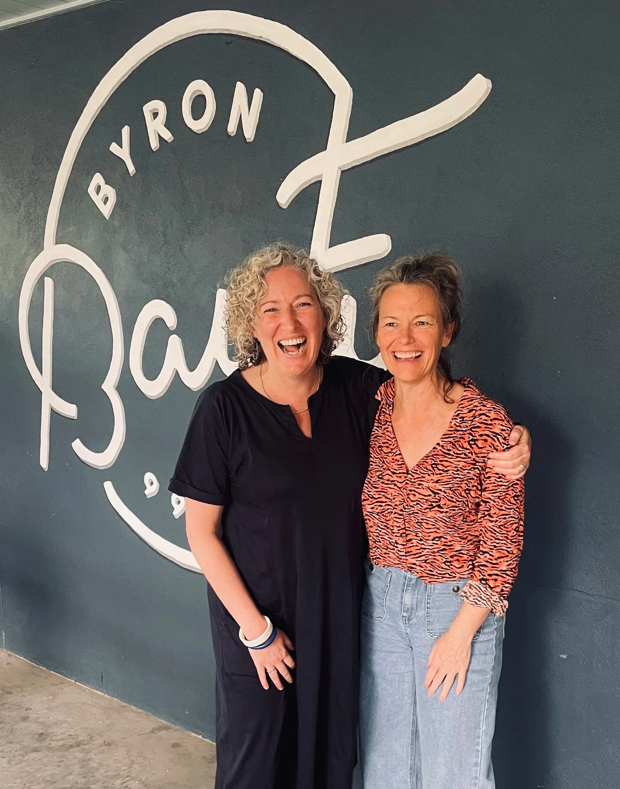 Bonita on the left and Emma on the right hugging and laughing. They are standing in front of a wall that says Byron BayFM.