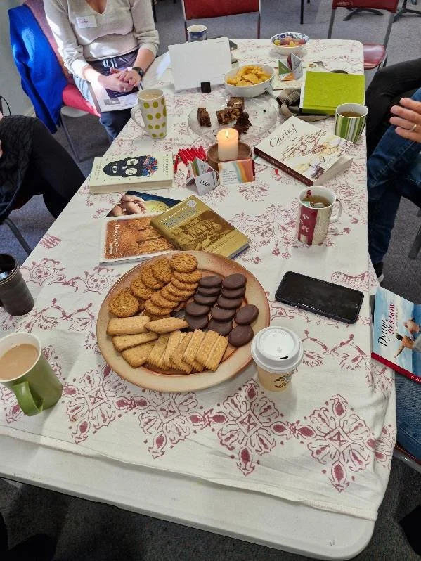 A table with a large plate of assorted cookies including chocolate-coated, rectangular, and round cookies, surrounded by coffee mugs, books, a lit candle, and snacks. People are gathered around, some holding papers, with chairs visible in the backgro