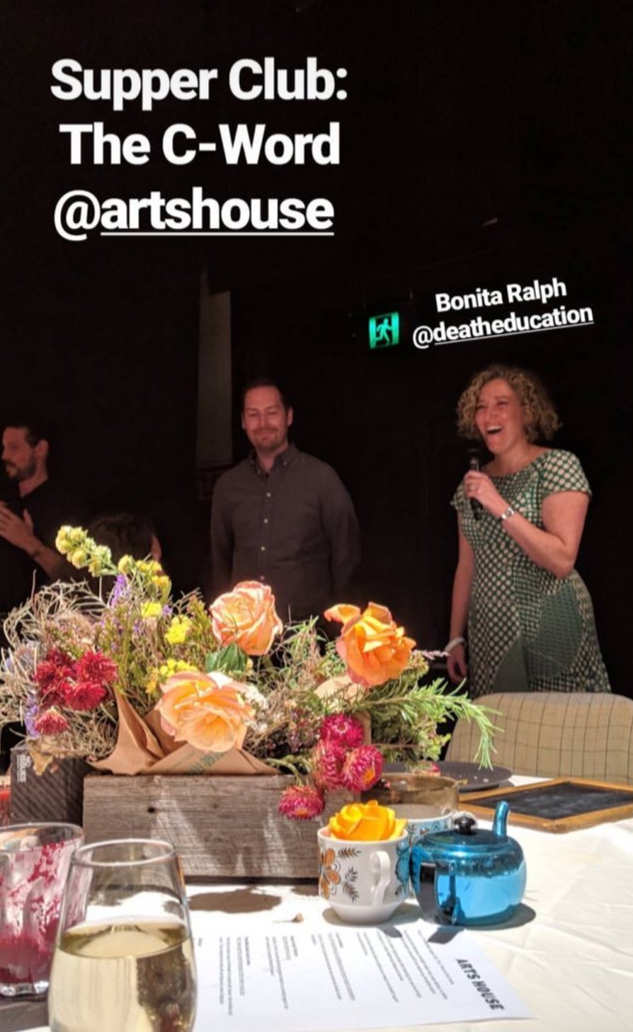 A woman speaking at a dinner event with floral centerpieces on the table, while two men stand behind in a dark room, with text overlay about the Supper Club event.