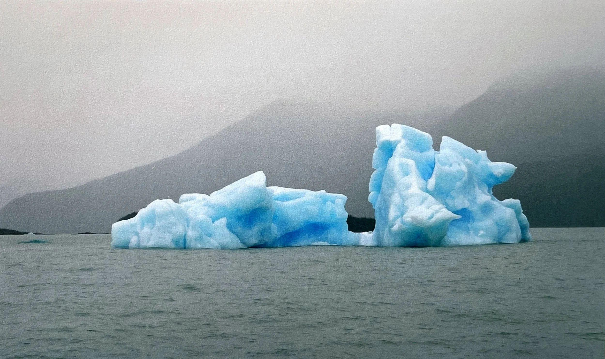 Two large icebergs floating in cold ocean water with mountains in the background, under an overcast sky. Representing that when we think about death, there is much more below this that what needs to be thought about on the surface.
