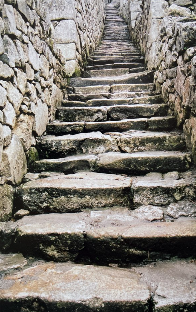 Old stone staircase with uneven steps, flanked by walls made of large stones, ascending upward. Image is from the Macchu Picchu historical site.