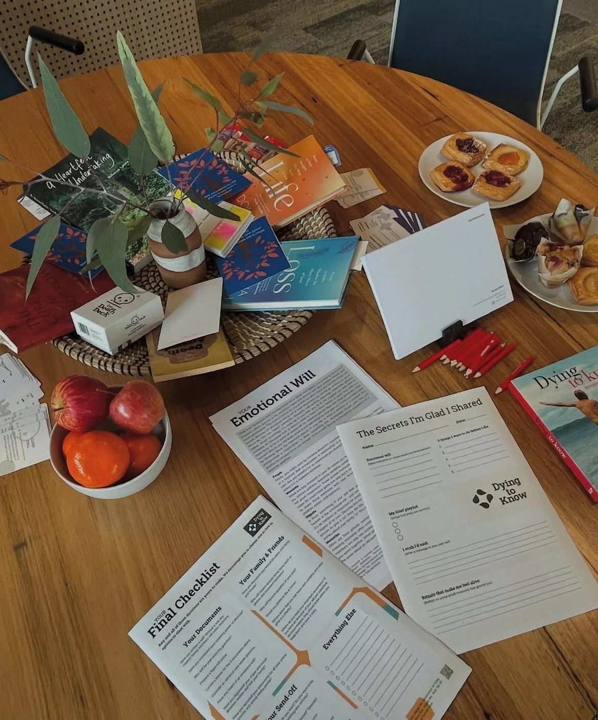 A wooden table with brochures, a bowl of apples and oranges, papers, a plate of pastries, and a potted plant.