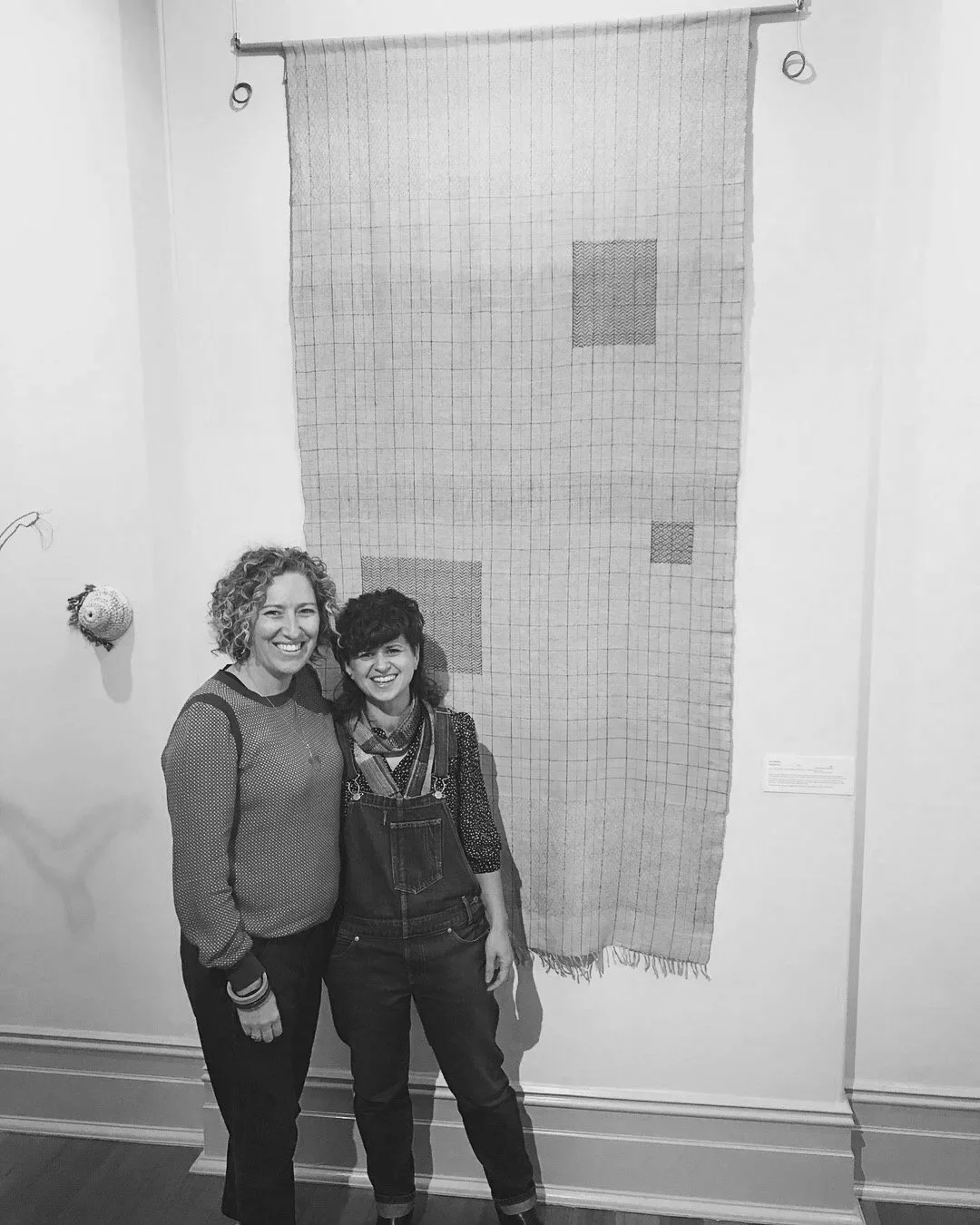 Two women smiling and standing close together in front of a hanging woven textile art with geometric patterns in a gallery or museum setting.