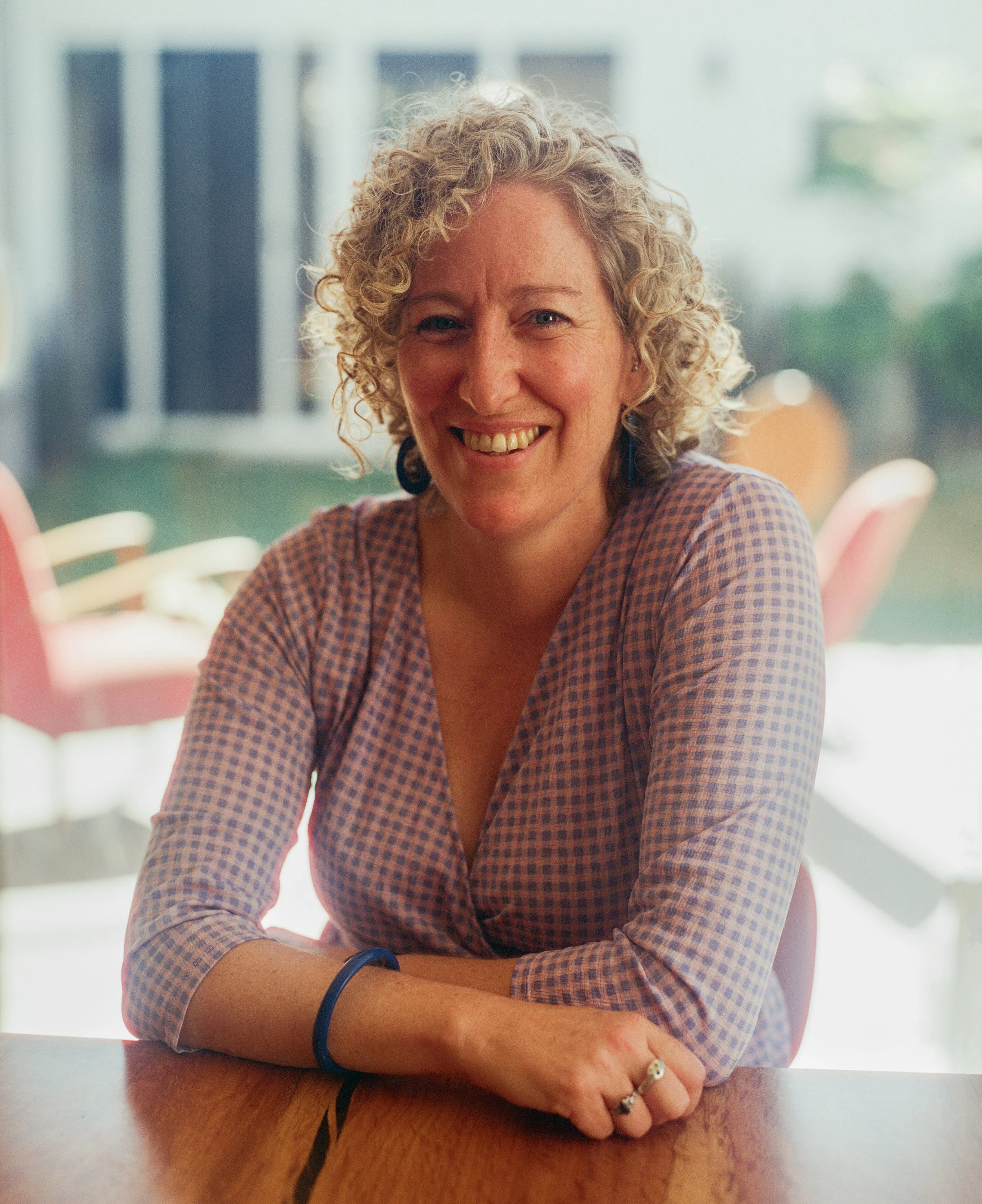 A woman with short curly blonde hair smiling at the camera, sitting at a wooden table in a bright room with large windows and pink chairs in the background.