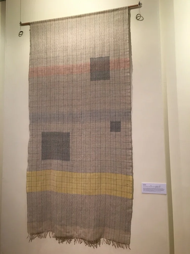 A textile wall hanging with a grid pattern, featuring squares and bands of color, including red, black, and yellow, displayed on a wall.