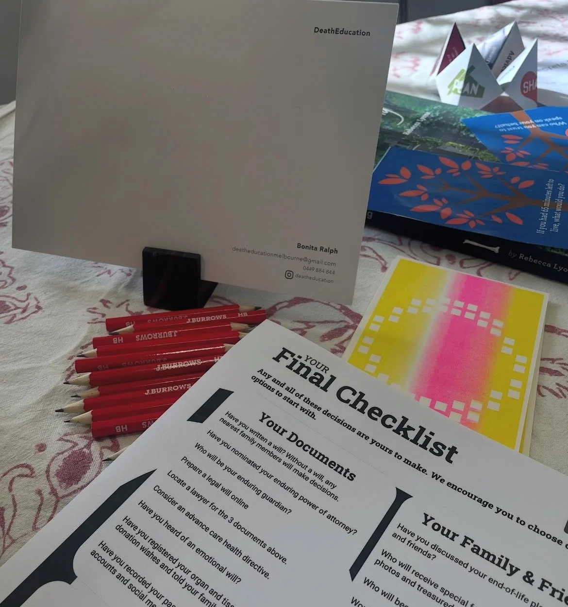 A table with various items including red pens labeled 'J. Burrows', a printed checklist titled 'YOUR Final Checklist', an open booklet titled 'Death Education' by Bonita Ralph, colorful flyers or brochures, and a patterned tablecloth.