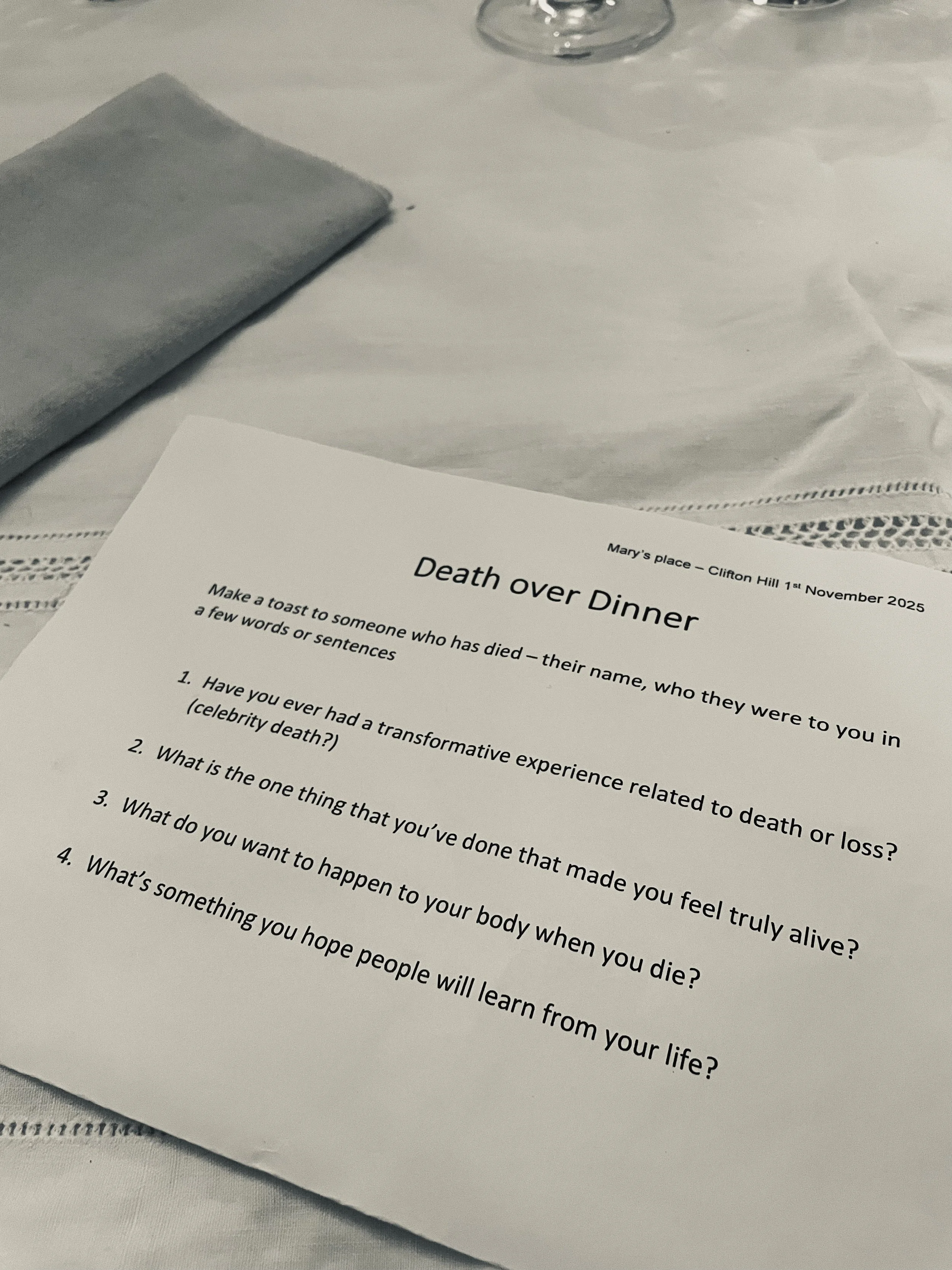 A printed worksheet titled 'Death over Dinner' on a table, with questions about death and life, with a napkin, a glass, and a tablecloth visible.