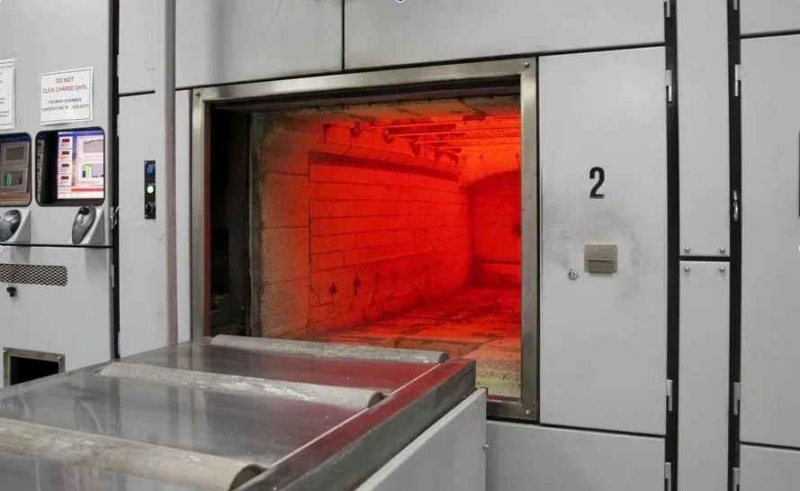 A cremator with door open. Glowing orange & red. There is a stainless stell trolley in front that has rollers