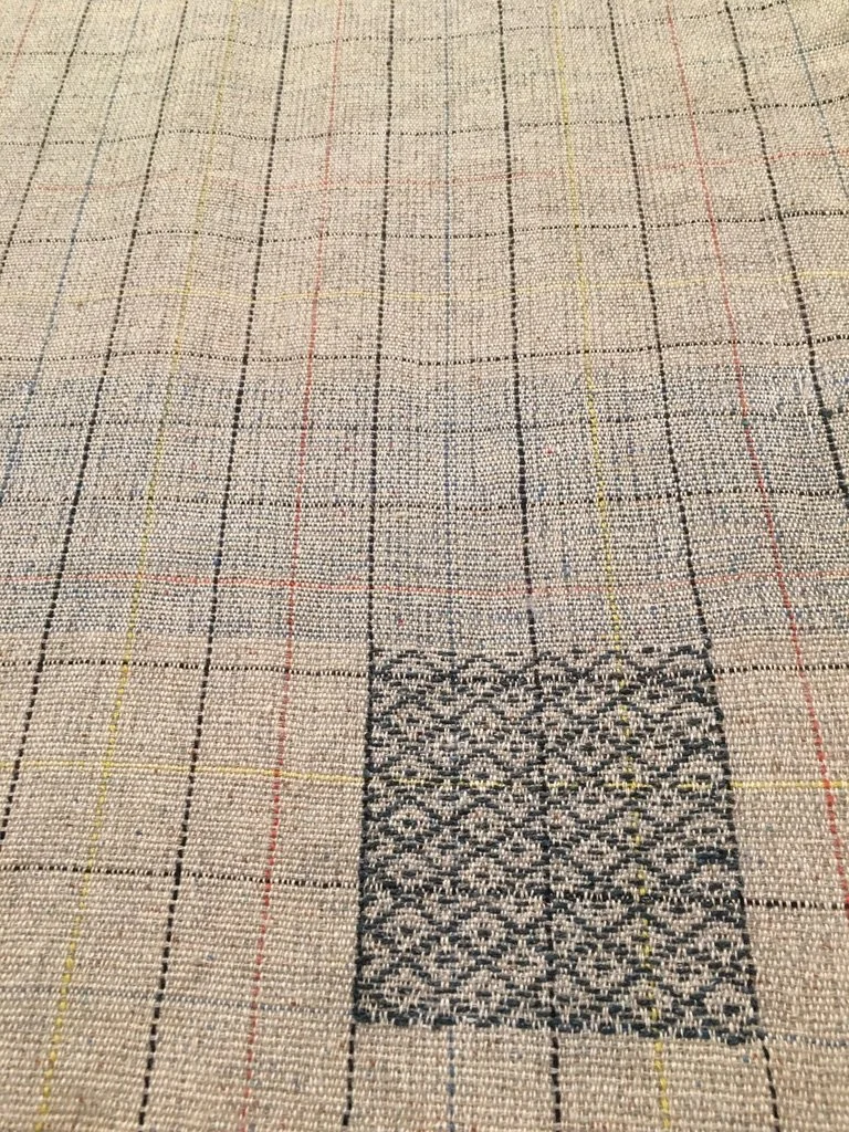 A woven fabric with a beige background, featuring a plaid pattern with black, red, and yellow lines and a decorative black and beige embroidered patch near the bottom.