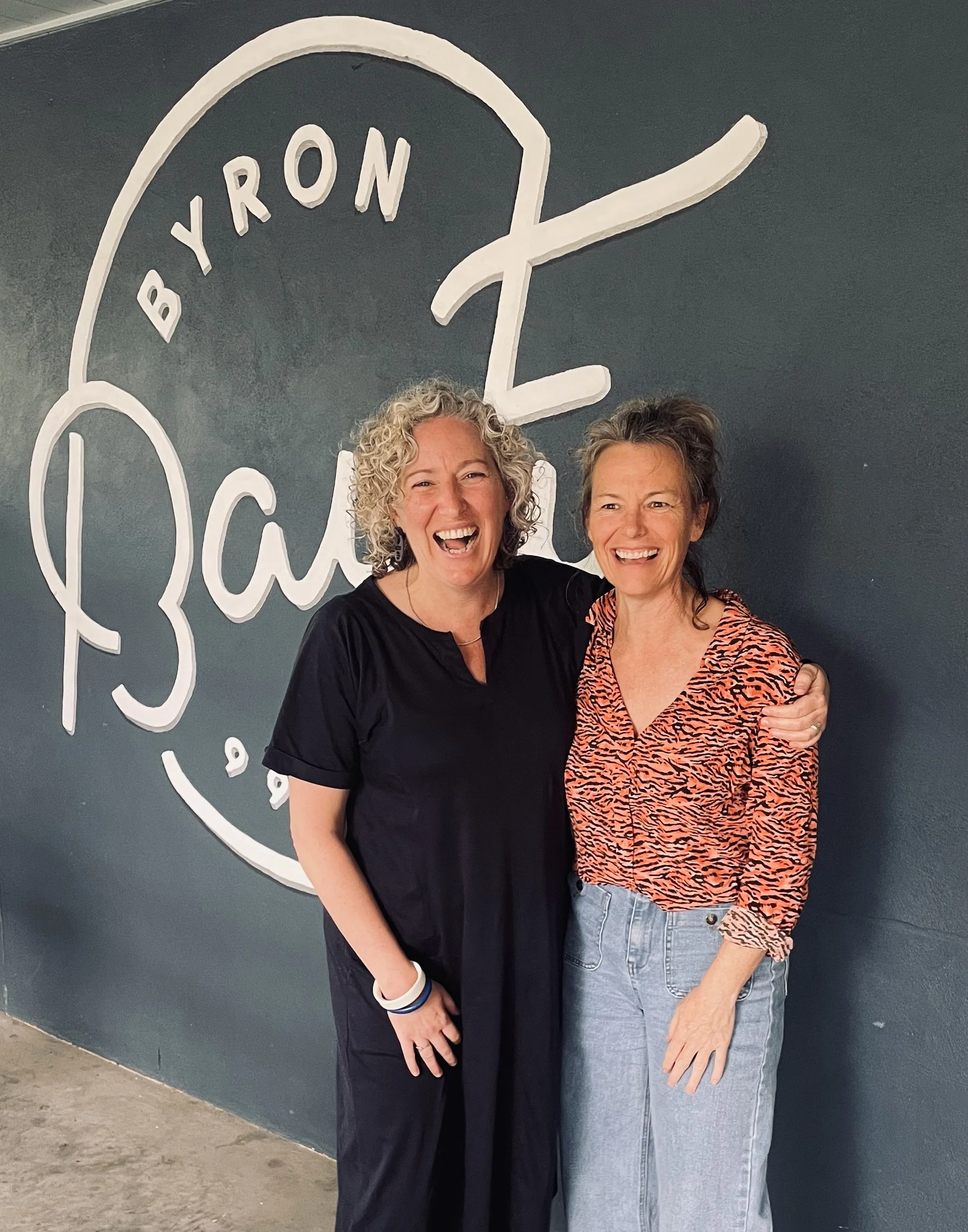 Two smiling women standing close together in front of a dark wall with a large white logo that says 'B yron Eat'.