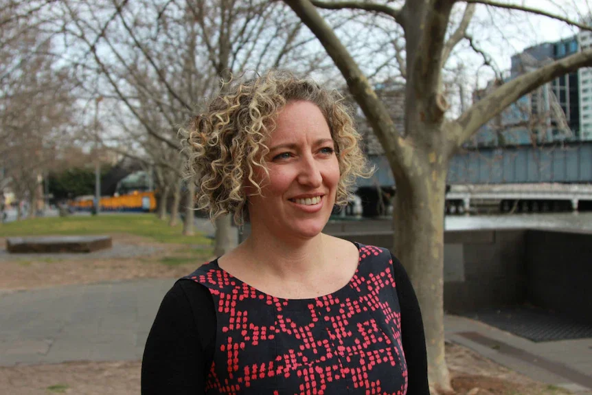 A woman with curly blonde hair smiling outdoors near a tree and a bridge, with a cityscape in the background.